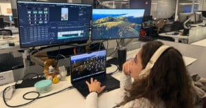 A person wearing headphones edits a video on a laptop connected to two large monitors at a desk. Various items, including a stuffed toy and tumbler, are on the desk in a modern office setting.