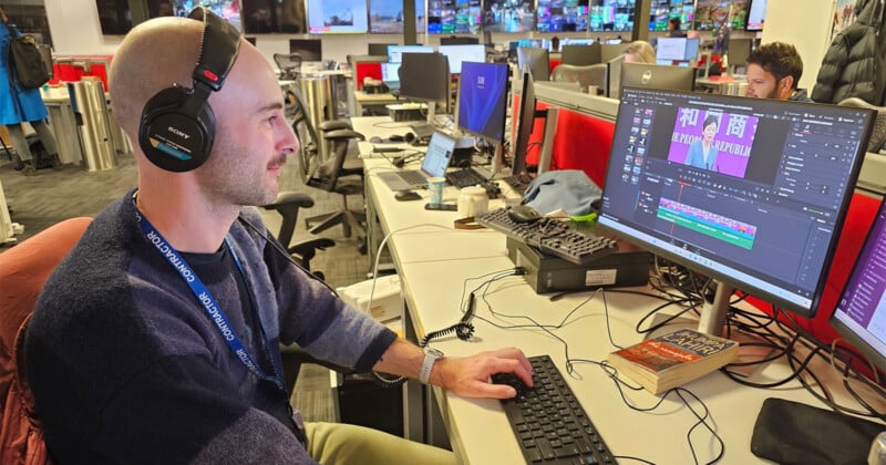 Un hombre con auriculares trabaja frente a una computadora en una oficina ocupada. El software de edición de vídeo estaba abierto en la pantalla y varios monitores y material de oficina estaban visibles sobre el escritorio. Otros trabajan en segundo plano.