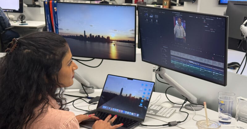 Una mujer sentada en su escritorio usando una computadora portátil conectada a dos monitores, uno que muestra una puesta de sol de la ciudad de Nueva York y el otro muestra un programa de edición de video que contiene imágenes de personas. Hay varios artículos de oficina y un vaso sobre la mesa.