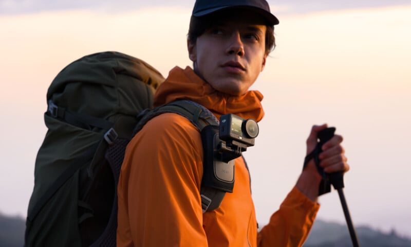 A person wearing an orange jacket and a cap hikes outdoors with a large backpack and trekking pole, and has an action camera mounted on their chest.