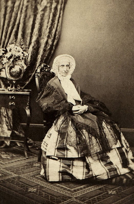 An elderly woman in Victorian-era clothing sits in a chair, wearing a bonnet and shawl. She holds a small book in her lap. Behind her is a table with a flower vase and a heavy draped curtain.