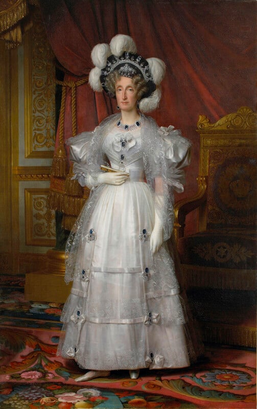 A woman in an elaborate white gown with lace and blue ribbon details stands beside a golden chair, wearing white gloves, jewelry, and a large black hat adorned with white plumes, set against a luxurious interior backdrop.