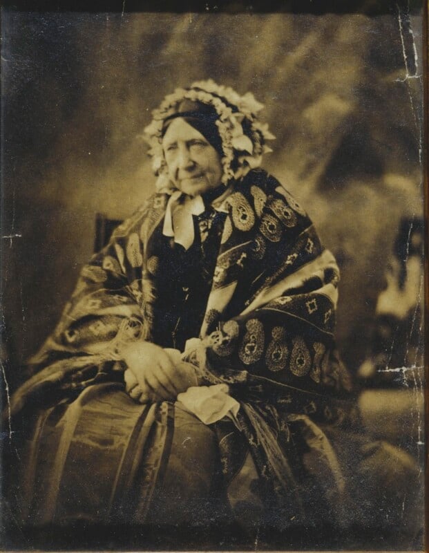 An elderly woman sits with her hands clasped, wearing a patterned shawl and a bonnet decorated with flowers, in a sepia-toned vintage photograph.