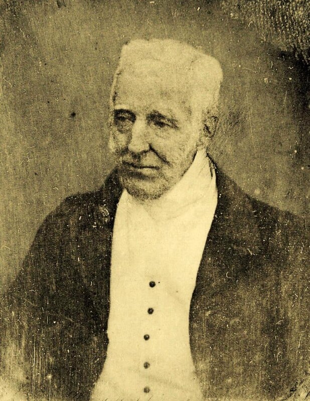 Sepia-toned portrait of an older man with short white hair, wearing a dark jacket over a high-collared white shirt with buttons, looking slightly to the side with a calm expression.