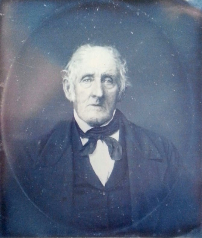 An older man with white hair, wearing a dark suit, white shirt, and a cravat, sits for a formal portrait. The photograph has a vintage, sepia-toned appearance, suggesting it is from the 19th century.