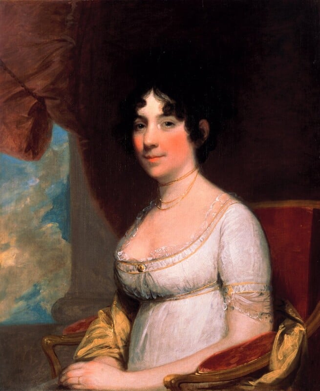 A woman with dark curly hair wears a white empire-waist dress and gold jewelry, seated on a red chair. She poses indoors beside a window with a red curtain and a view of blue sky and clouds.