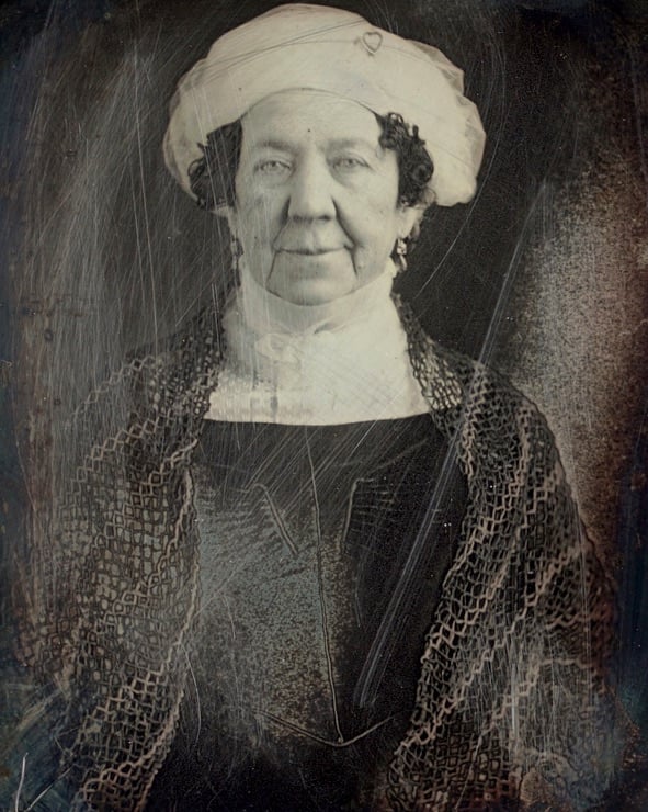 A vintage, scratched portrait of an older woman wearing a white bonnet, white scarf, and dark dress with a patterned shawl, looking at the camera with a slight smile.