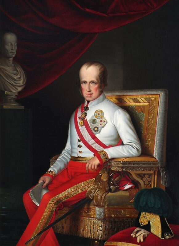 A man in a decorated military uniform with medals and a red sash sits on a golden ornate chair, holding a document. There is a red draped curtain and a bust in the background, with a plumed hat on a table beside him.