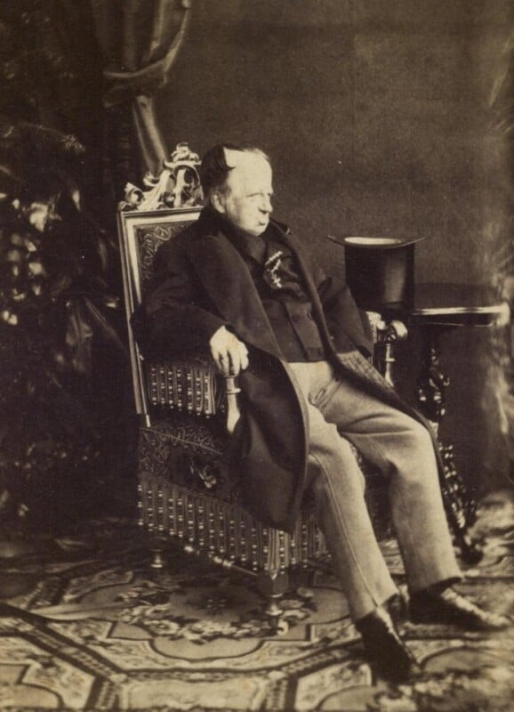 An older man with light hair and sideburns sits on an ornate chair, wearing a dark coat and light trousers. A top hat rests on a small table beside him, and the setting includes patterned carpet and draped fabric.
