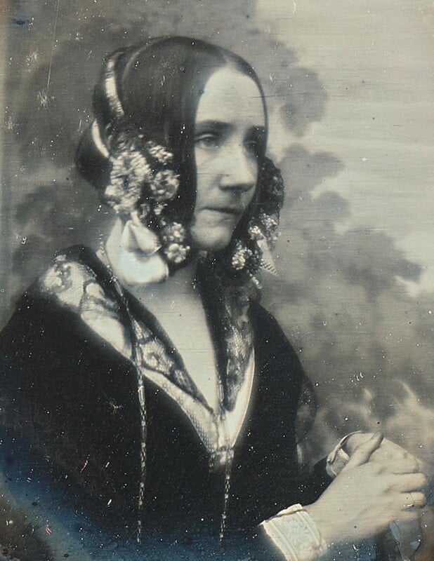 A black-and-white vintage portrait of a woman with dark hair arranged in elaborate curls, wearing a decorated dress with lace and floral adornments, standing with her hands clasped in front of a blurred background.