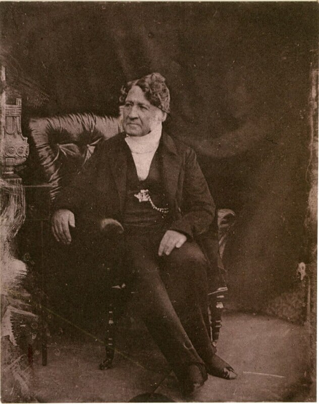 An older man with wavy hair sits on a cushioned chair, dressed in formal 19th-century attire including a dark suit, white cravat, and watch chain, facing slightly to the side against a dark backdrop.