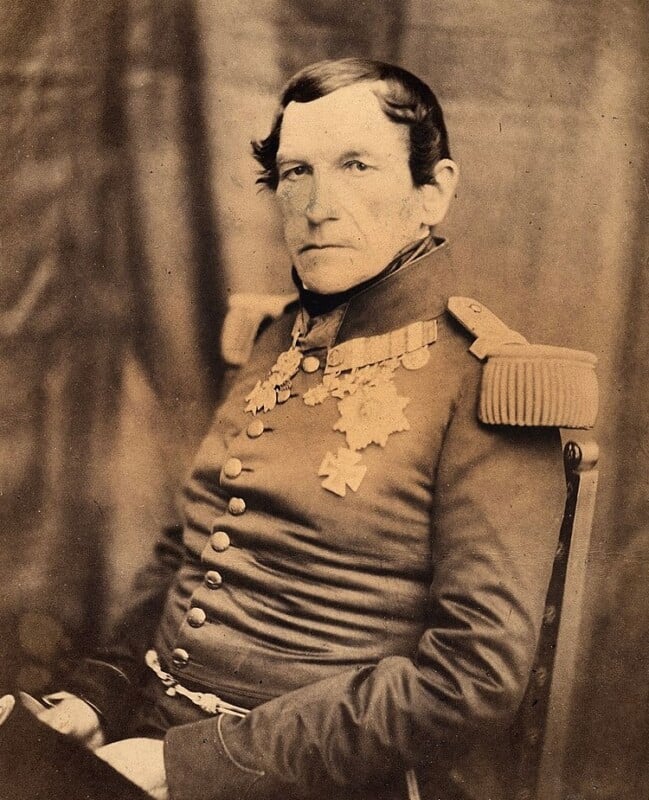 Sepia-toned portrait of a stern-faced man in a decorated military uniform with epaulets, medals, and a double row of buttons, seated against a plain backdrop.