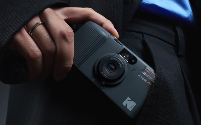 A person wearing a dark suit and a silver ring on their finger is holding a black Kodak digital camera, partially tucked into their pocket.
