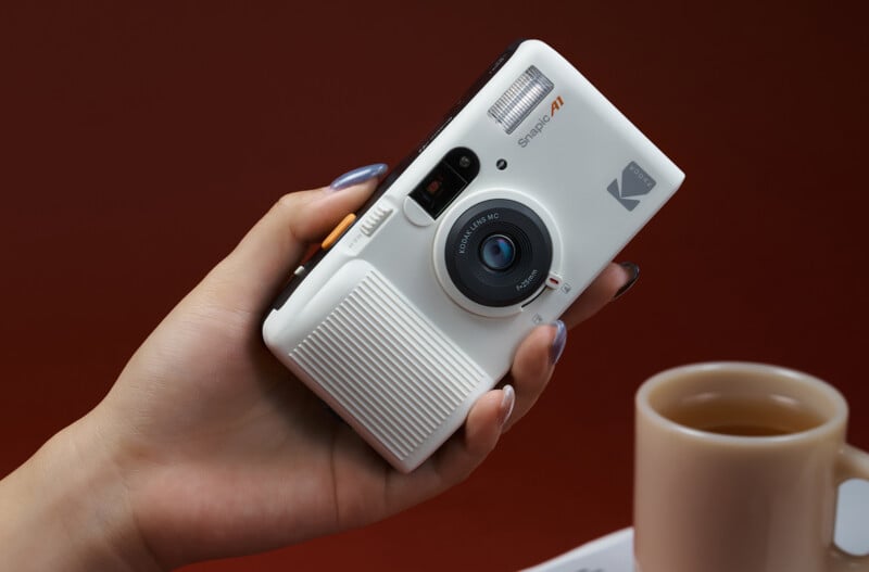 A hand holding a white Kodak Snap A1 camera over a table with a beige cup of tea in the background, against a red backdrop.