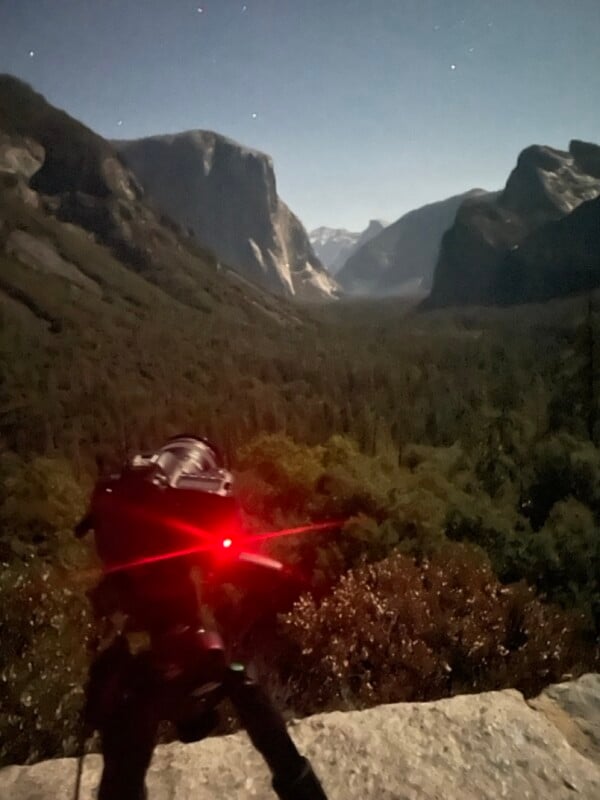 Una cámara con una luz roja montada en un trípode apunta a un pintoresco valle con imponentes acantilados y densos bosques bajo un cielo nocturno estrellado.
