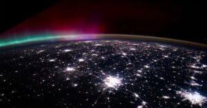 A view of Earth from space at night, showing bright city lights and a colorful aurora with green and pink hues in the atmosphere above the horizon.
