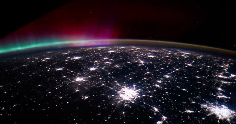A view of Earth from space at night, showing bright city lights and a colorful aurora with green and pink hues in the atmosphere above the horizon.