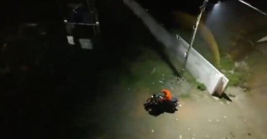 A person wearing an orange top sits on a motorcycle at night near a white wall and a streetlight, with dark surroundings and patches of grass visible on the ground.