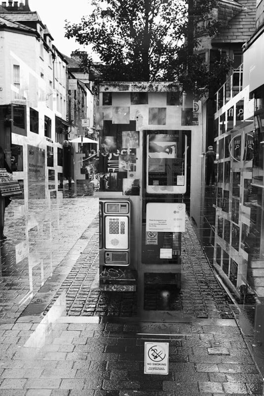 A black-and-white photo of a payphone booth on a wet, cobblestone street lined with buildings and trees, surrounded by posters and reflections in glass panels, on a rainy day.