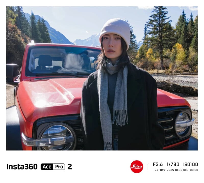 Un hombre que lleva un gorro gris claro, una bufanda gris y un abrigo oscuro se encuentra frente a un SUV rojo contra un paisaje de bosque montañoso bajo un cielo azul claro.