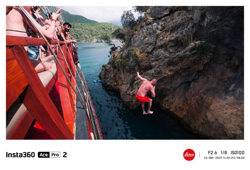 Un hombre con bañador rojo salta de un barco al agua mientras los espectadores observan y toman fotografías; acantilados rocosos y árboles verdes rodean la escena en un día soleado.