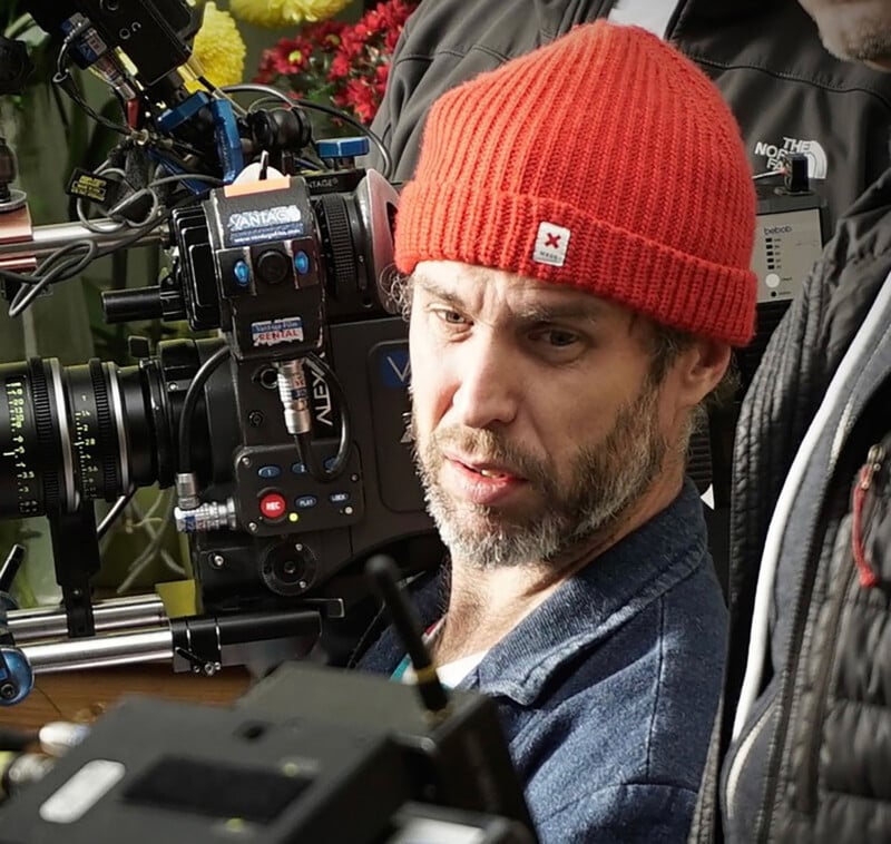 Un hombre con barba corta y gorro de punto rojo maneja una cámara de vídeo profesional y está concentrado en su trabajo. Hay varios accesorios y etiquetas en la cámara con personas borrosas y flores de fondo.