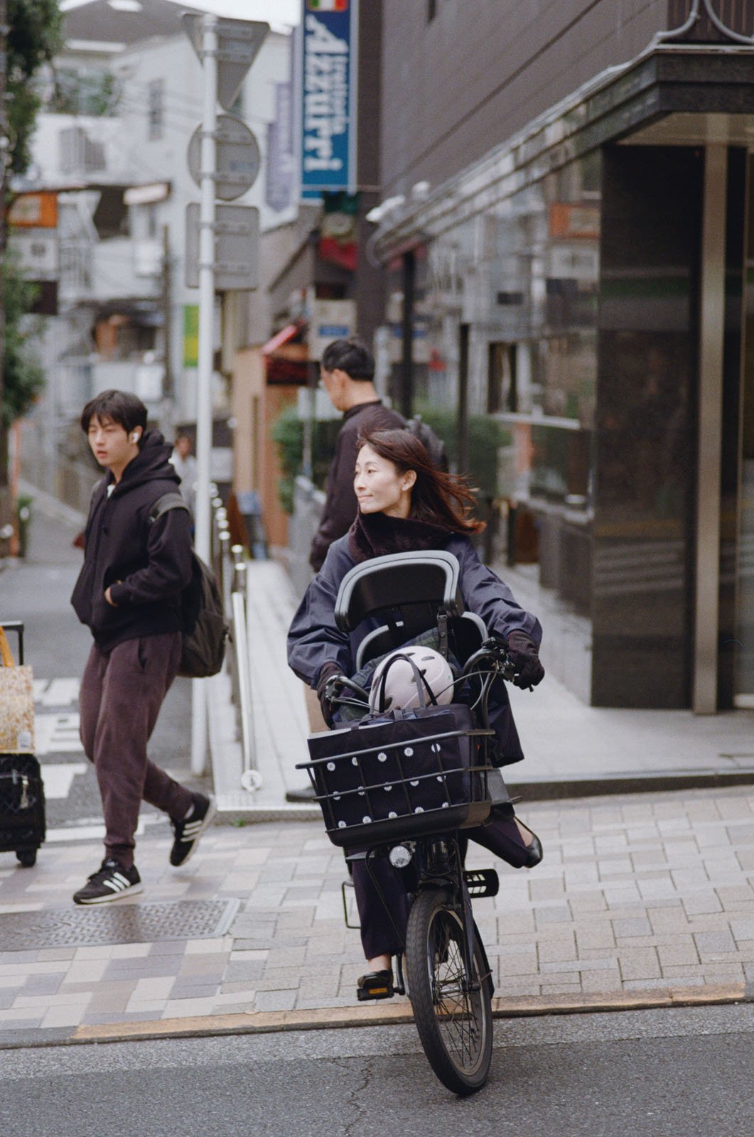 A woman rides a bicycle with a child seat on a city street. Two pedestrians, dressed in dark clothing, walk nearby. Urban buildings and signs are visible in the background.