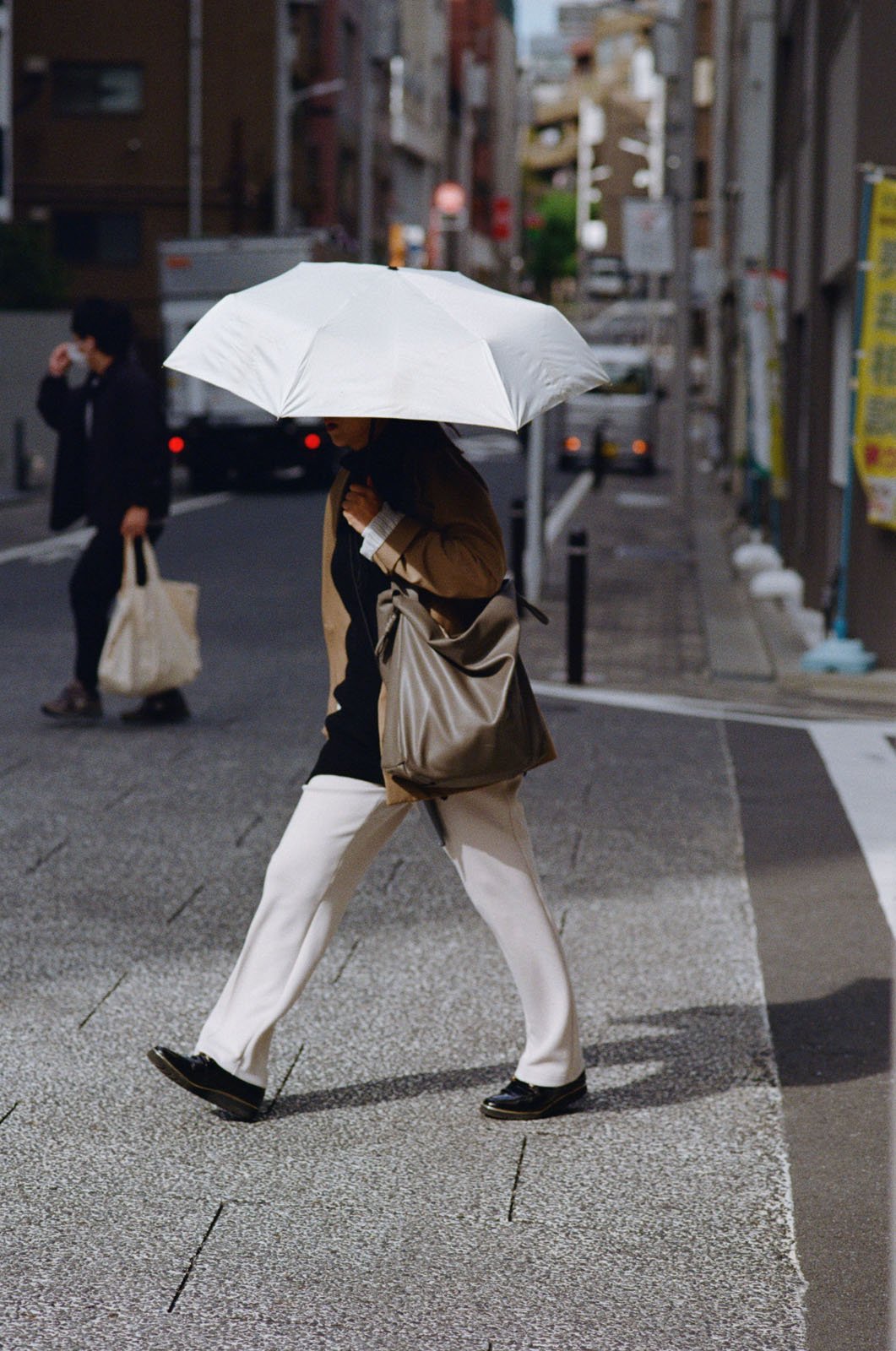 A person walks across a city street holding a large white umbrella. They wear white pants, a tan coat, and carry a gray bag. The urban background shows buildings, signs, and another person with a shopping bag.