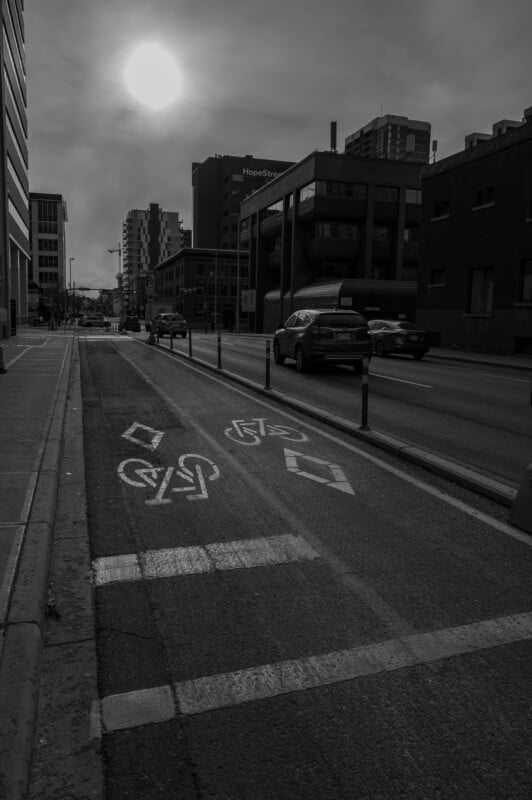 Una fotografía en blanco y negro de automóviles circulando por una calle de la ciudad, un carril bici separado marcado con símbolos, rodeado de edificios altos a ambos lados y la luz del sol brillando a través de un cielo nublado.