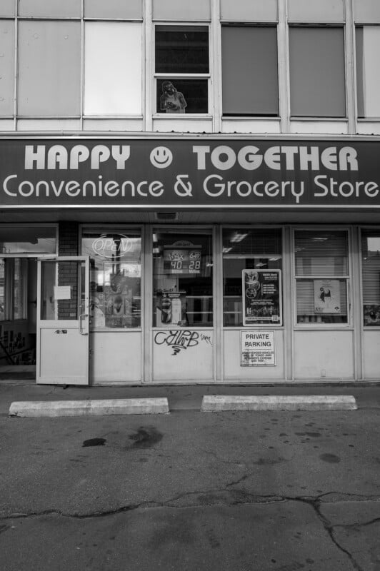 foto en blanco y negro "Tienda de comestibles Happy Together" La tienda tiene un letrero, carteles en las ventanas y dos espacios de estacionamiento vacíos en el frente. La carita sonriente es parte del letrero de la tienda.