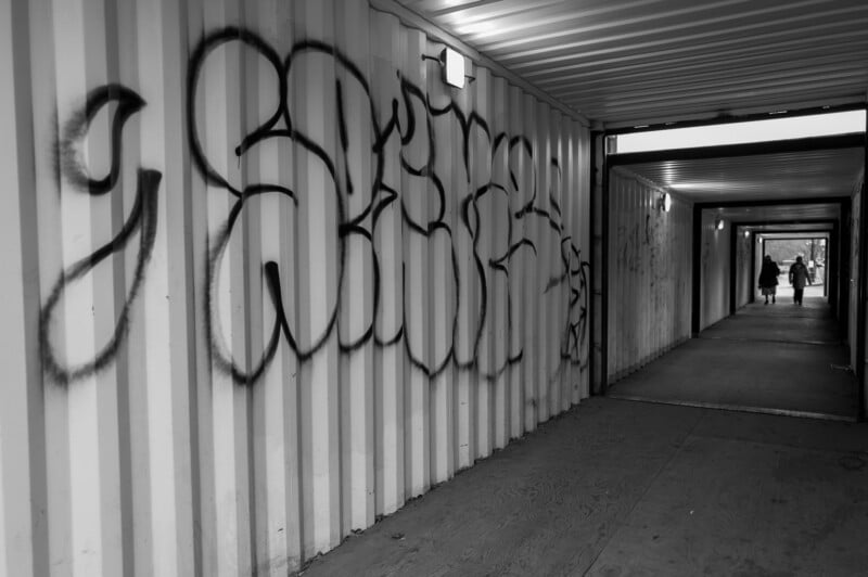 Una fotografía en blanco y negro de un pasillo largo y estrecho con graffiti en la pared izquierda y dos personas caminando a lo lejos. El pasillo está iluminado por luces de techo.