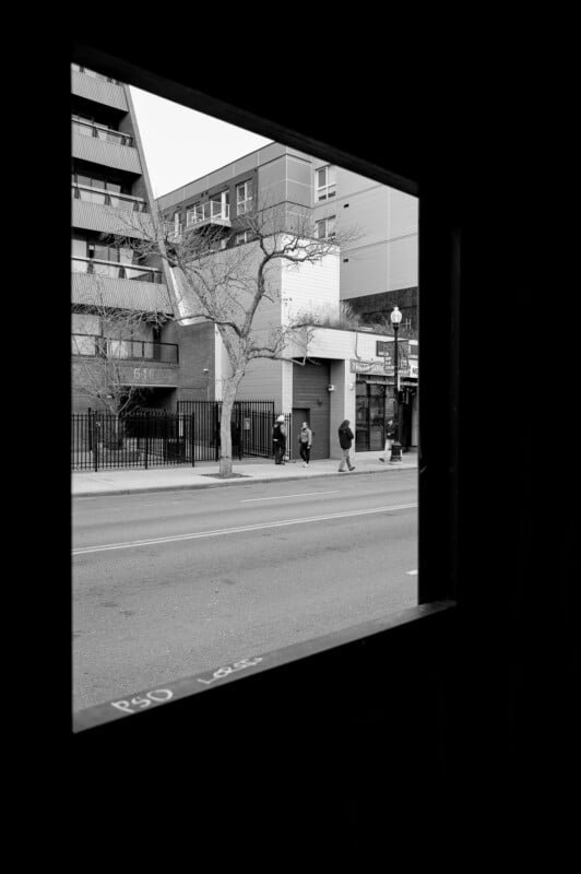 La escena callejera de la ciudad está enmarcada por una ventana oscura. Cuatro personas caminaban por la acera junto a edificios y árboles desnudos. Las fotos están en blanco y negro.