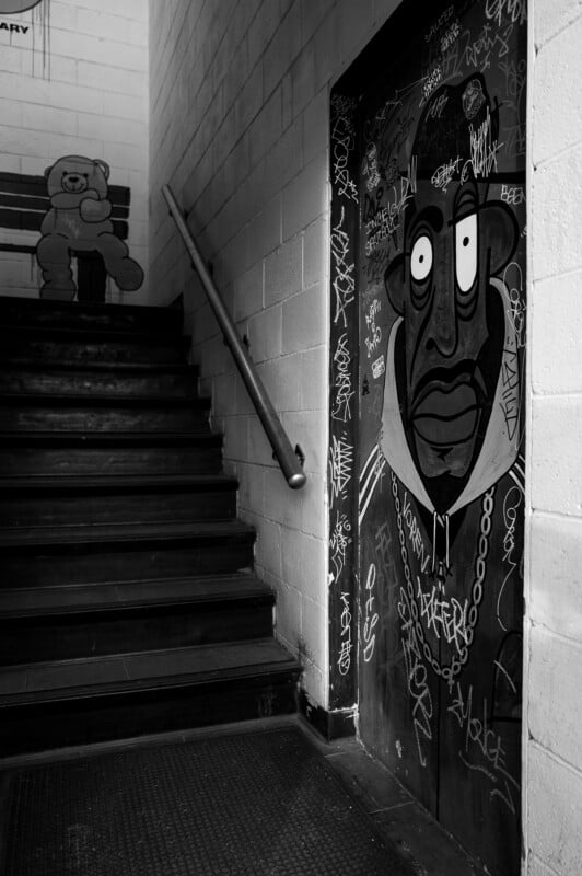 Una fotografía en blanco y negro de una escalera con graffiti; una cara de dibujos animados con una gran cadena dibujada en la puerta y un oso sentado en un banco en lo alto de las escaleras. Las paredes y puertas estaban cubiertas de etiquetas.