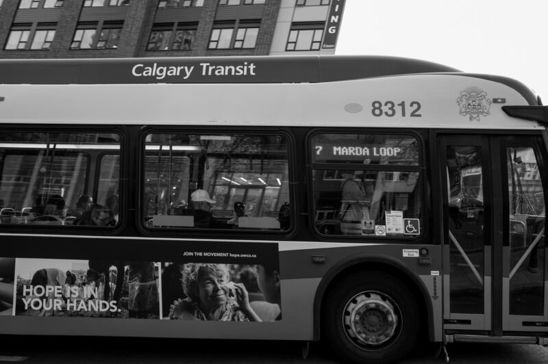 Fotografía en blanco y negro de un autobús de Calgary Transit, ruta 7 con destino a Malda Loop. El anuncio al lado dice "La esperanza está en tus manos" A su lado hay una anciana sonriente. Al fondo se ven claramente los edificios de la ciudad.