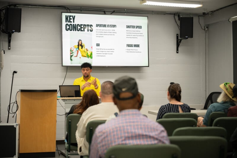 A man in a yellow shirt teaches a photography class in a small classroom. He stands in front of a screen displaying key concepts while students sit and listen attentively.