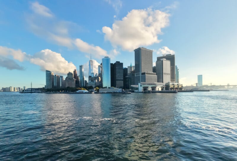 El horizonte de la ciudad de Nueva York se ve desde el agua, con imponentes rascacielos, cielo azul, nubes dispersas y reflejos brillantes en el río.