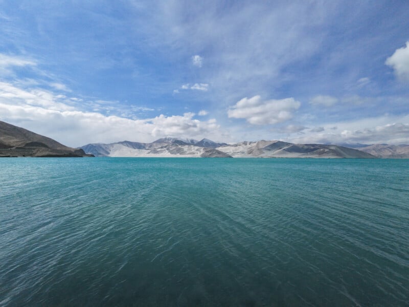 El vasto lago color turquesa tiene suaves ondas, rodeado de lejanas montañas cubiertas de nieve bajo un cielo azul y nubes blancas.