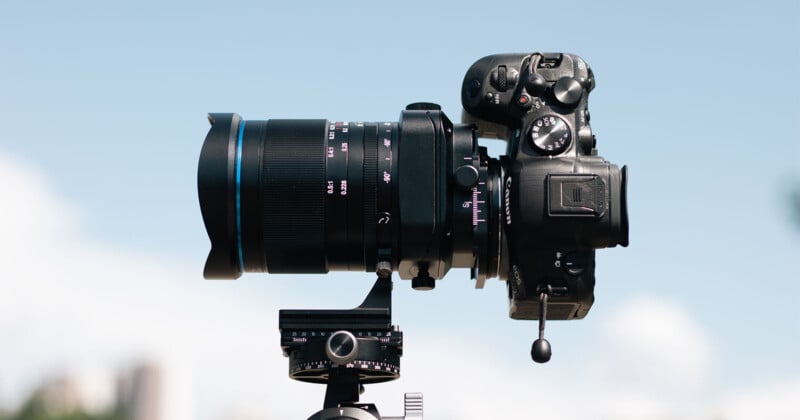 A professional DSLR camera with a large lens is mounted on a tripod against a clear sky background. The camera is viewed from the side, showing detailed buttons and dials.
