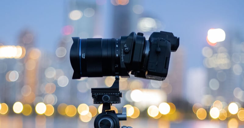 A black DSLR camera with a large lens is mounted on a tripod, positioned against a blurred cityscape background with colorful bokeh lights.