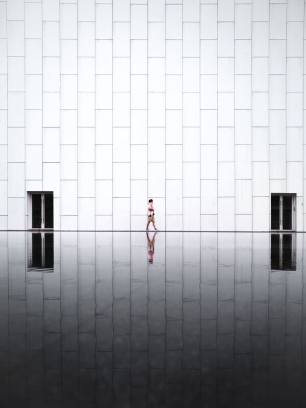 A person walks past a large white tiled wall with two doorways, reflected in a still, dark pool of water below. The minimalist scene emphasizes symmetry and scale.