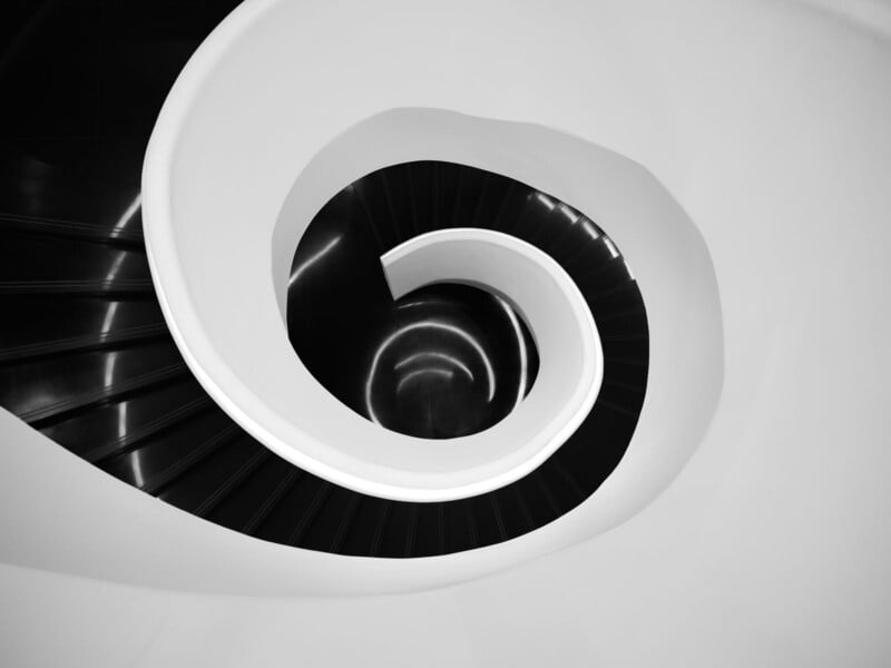 A black and white photo of a modern spiral staircase, viewed from above, showing swirling steps and smooth, curved lines forming a circular pattern.