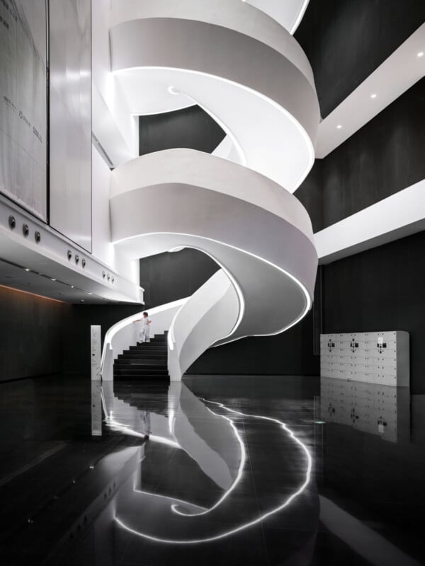 A modern interior with a large, white spiral staircase featuring a glowing edge, reflected on the shiny black floor. A person in white stands on the stairs beneath high ceilings and dark walls.
