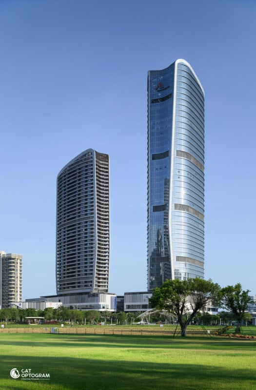 Two modern, glass-covered skyscrapers rise side by side against a clear blue sky, with green lawns and trees in the foreground and smaller buildings nearby.