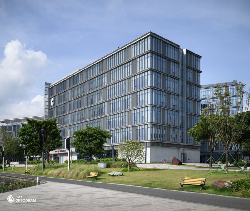 A modern, multi-story glass office building with trees, benches, and a walkway in the foreground on a sunny day. The building features large windows and a minimalist design.