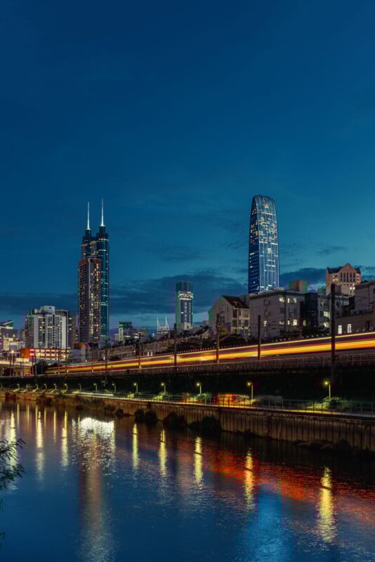 City skyline at dusk with illuminated skyscrapers, a river reflecting colorful lights, and a train passing by, leaving light trails against the deep blue evening sky.