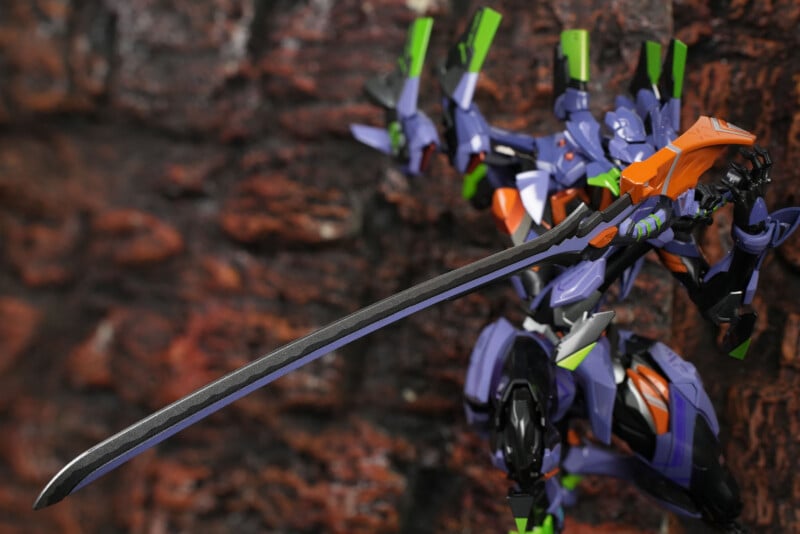 A detailed action figure of Evangelion Unit-01 from "Neon Genesis Evangelion" wields a large sword, posed dynamically against a textured brick wall background.