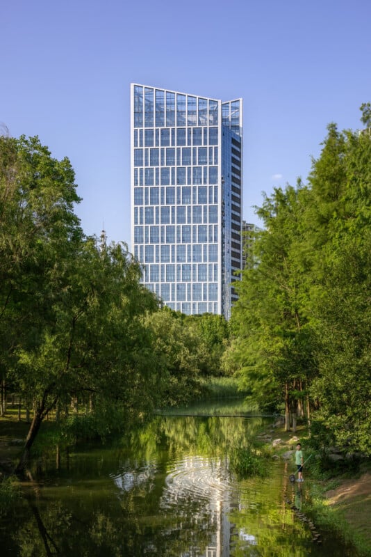 A modern glass skyscraper with a geometric facade stands behind lush green trees, reflected in a calm pond below. A person in a hat stands near the water’s edge, fishing amidst the greenery under a clear blue sky.