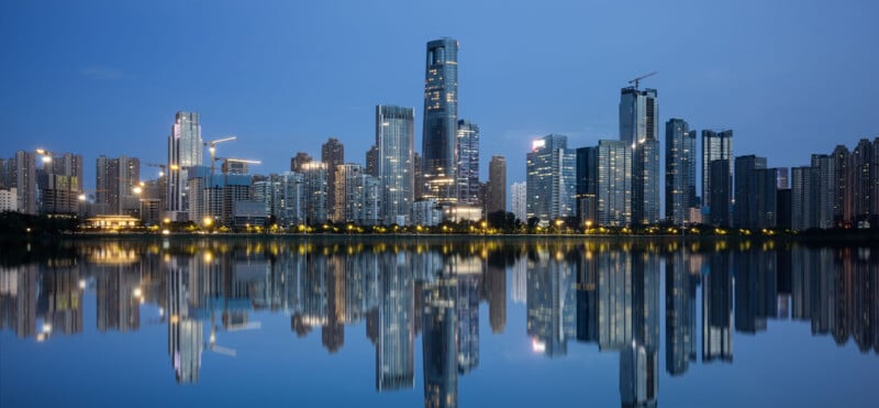 A modern city skyline with tall skyscrapers is reflected in a calm body of water at dusk, with building lights and a clear blue sky creating a serene urban scene.