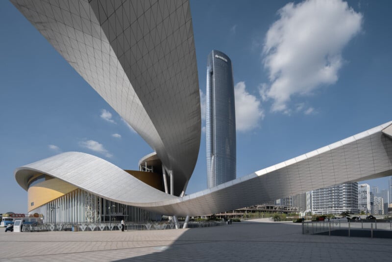 A futuristic building with sweeping, curved metal and glass roofs stands in front of a tall, cylindrical skyscraper under a blue sky with scattered clouds. Modern city buildings are visible in the background.