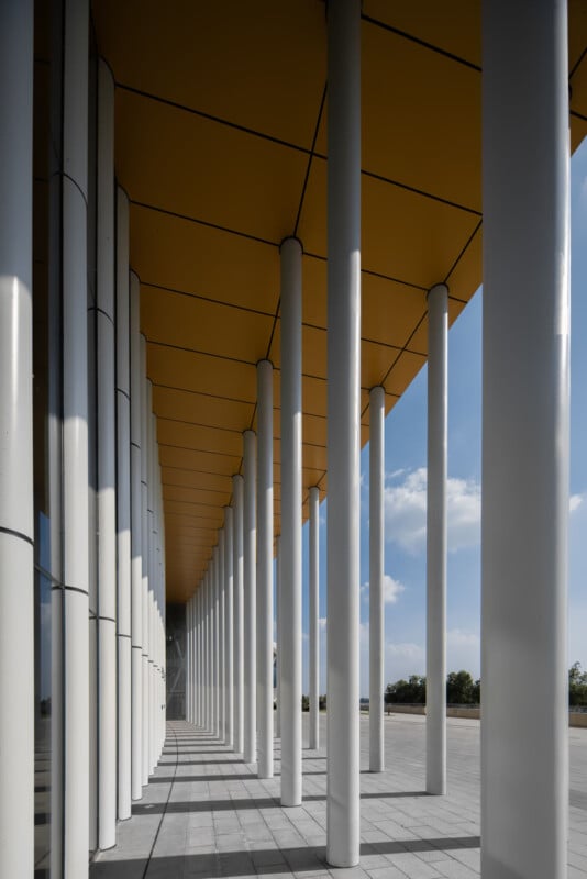 A modern building with tall white columns and a yellow ceiling, casting shadows on the gray tiled ground. The columns create a geometric pattern, and blue sky with clouds is visible in the background.